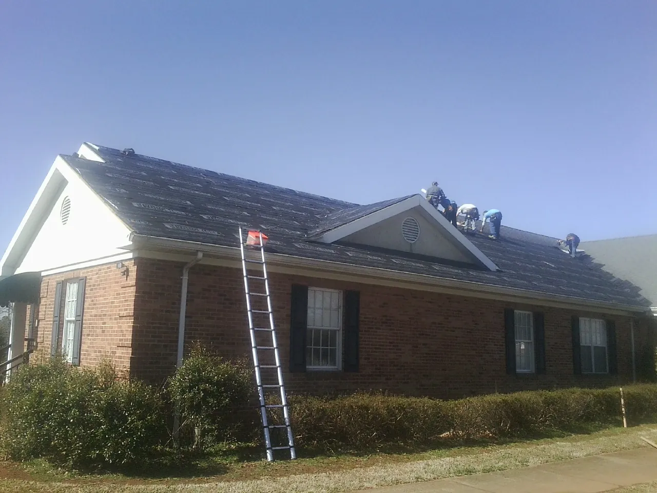 Crew tearing shingles off a brick ranch roof with a ladder set — Metal Roof Repair work by our crew in Cotuit